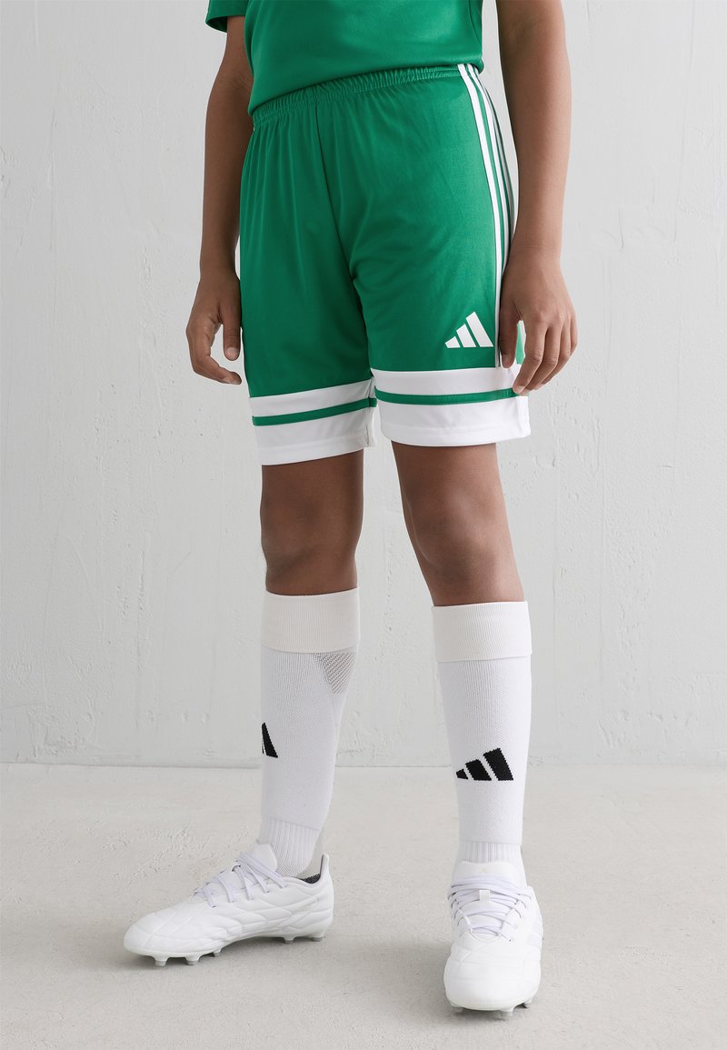 Barn som bär gröna Adidas sportshorts, vita knähöga strumpor med svart logotyp och vita fotbollsskor som står på en grå golv.