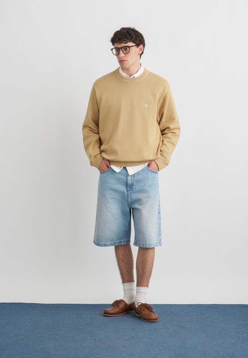 Jeune homme portant des lunettes, pull beige sur chemise blanche, short en denim bleu clair, chaussettes blanches et mocassins marron, debout sur un tapis bleu.