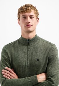 Groene ritstrui met een textuurpatroon, voorzien van een hoge kraag en een klein hexagonaal logo op de linkerkant van de borst.