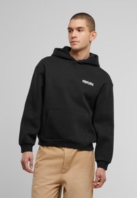 Schwarzer Baumwoll-Hoodie mit einer Vorderseite-Tasche und weißem Logo auf der Brust. Hat eine Kapuze mit Kordelzug und gerippten Bündchen; getragen mit beigen Hosen.