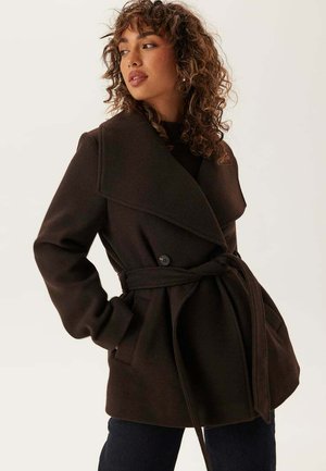 Kvinna med lockigt hår som bär en mörkbrun kappa med bälte och bred krage, händerna i fickorna, tittar åt sidan mot en enkel bakgrund.