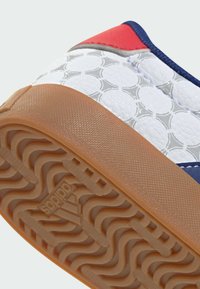 Baskets blanches avec motifs circulaires, semelle marron texturée avec logo Adidas, languette rouge au talon et bordure bleue.