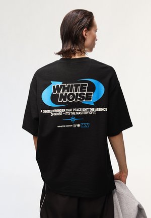 Person with wet hair wearing black T-shirt with blue arrows and text "WHITE NOISE" and a quote about peace and noise on the back.