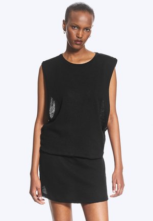 Woman with short hair wearing a black sleeveless dress with padded shoulders, standing against a white background with a neutral expression.