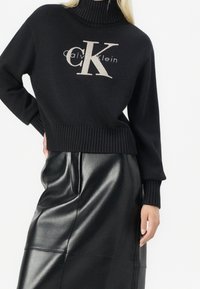 Pull à col roulé en maille noire avec un grand logo "CK" beige et le texte "Calvin Klein". Associé à une jupe en cuir noire, créant un look élégant.