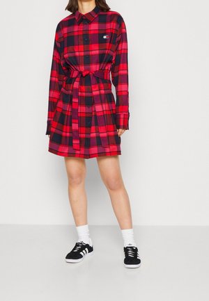 Femme portant une robe-chemise à carreaux rouges et noirs avec une ceinture, assortie de chaussettes blanches et de baskets Adidas noires à rayures blanches.