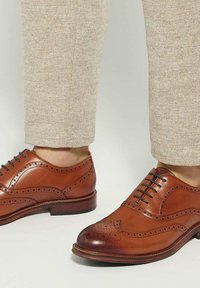 Chaussures en cuir marron à lacets avec des détails perforés, un bout arrondi et une finition lisse, associées à un pantalon texturé beige clair.