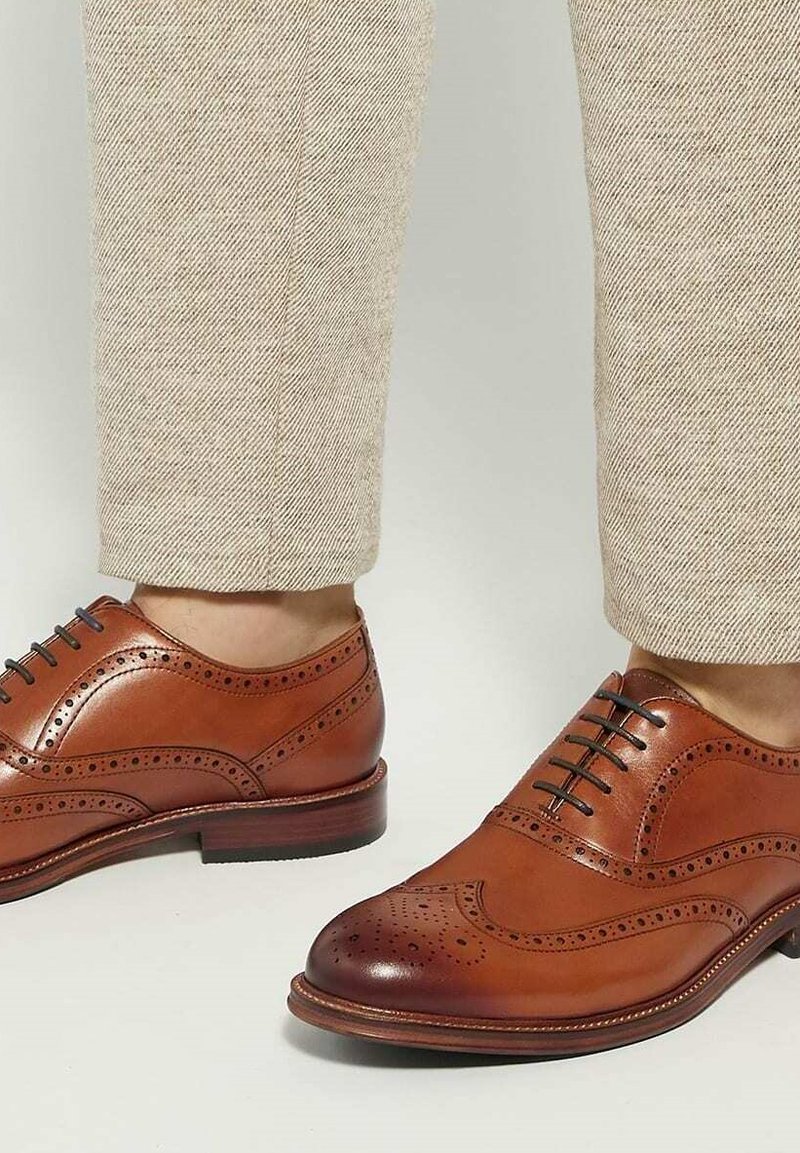 Chaussures en cuir marron à lacets avec des détails perforés, un bout arrondi et une finition lisse, associées à un pantalon texturé beige clair.