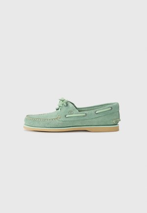 Chaussure bateau en suede vert avec un avant rond, un laçage à l'avant et une semelle en caoutchouc contrastante beige. Présente des coutures décoratives et des œillets en métal.