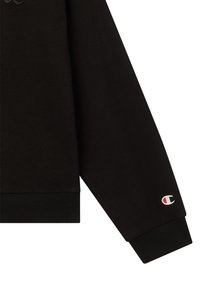 Manica di felpa nera con polsino a costine, dotata di piccolo logo Champion bianco, rosso e nero vicino al polso.