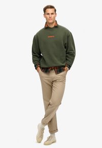 Groene fleece sweatshirt met een oranje logo, gelaagd over een rood-zwart geruit shirt. Beige broeken en lichtgekleurde sneakers maken de look compleet.