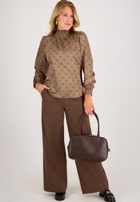 Bruine blouse met een hoog smock-kraag en lange mouwen, gecombineerd met brede bruine broeken. Houdt een donkerbruine handtas vast.