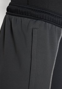 Tamsiai pilki sportiniai kelnių modeliai pasižymi tekstūruota medžiaga, elastinga juosmens juosta ir šoniniu kiemeliu su kontrastingais siuvinėjimo akcentais.