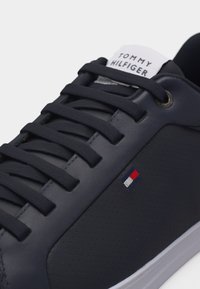 Κοντινό πλάνο σε ναυτικό μπλε αθλητικό παπούτσι με μαύρα κορδόνια, διάτρητη πλαϊνή πλευρά, ετικέτα λογότυπου Tommy Hilfiger και ετικέτα με εμπορικό σήμα στη γλώσσα.