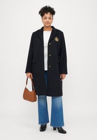 Fekete kabát hajtókával, arany gombdíszítéssel és emblémával. Fakó kék farmernadrággal és barna kézitáskával párosítva. Fekete magassarkú csizmák.