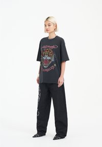 Svart oversized t-shirt med en färgglad tiger grafik och text, i kombination med vida svarta byxor med en vit design, och spetsiga svarta skor.