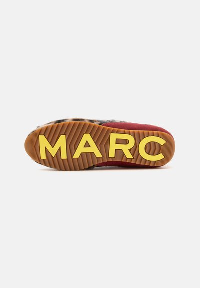 Semelle de chaussure avec de grosses lettres jaunes formant "MARC" sur une semelle en caoutchouc brun à rainures, avec des détails rouges et en fourrure sur la partie supérieure de la chaussure.