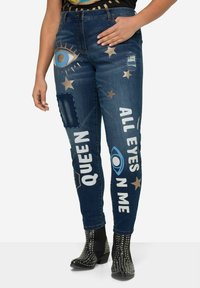 Jeans attillati in denim blu con grafiche decorative metalliche in oro e blu di occhi e stelle, toppe strappate, e scritte che recitano "QUEEN" e "ALL EYES ON ME."