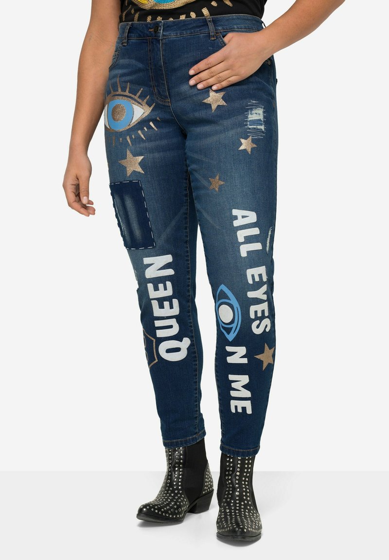 Jeans attillati in denim blu con grafiche decorative metalliche in oro e blu di occhi e stelle, toppe strappate, e scritte che recitano "QUEEN" e "ALL EYES ON ME."