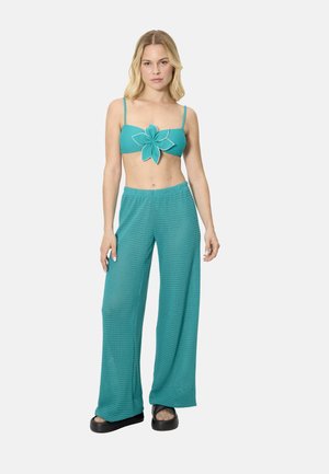 Mujer con pantalones anchos de pierna ancha en textura color teal y un top corto a juego con un gran detalle de flor, posando frente a un fondo liso.