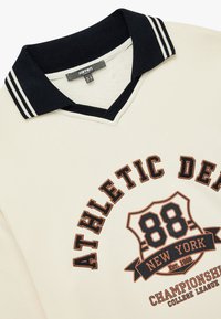 Beżowy sweter z granatowym kołnierzykiem, z białymi paskami. Zawiera grafikę z wyszytym napisem "ATHLETIC DEPT 88 NEW YORK" w ciemnym brązie.