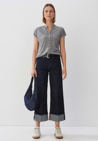 Camicia grigia a maniche corte con bottoni, jeans larghi di colore scuro con orli arrotolati, sneakers bianche e una borsa a tracolla blu navy.