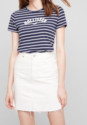 Kobieta ubrana w granatowo-biały pasiasty t-shirt Hollister w połączeniu z białą jeansową spódnicą z wysokim stanem i postrzępionym dołem.