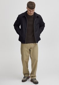 Sort zip-up jakke med spiss krage, kombinert med en brun ribbestrikket genser og beige løse bukser, båret med grå og røde sneakers.