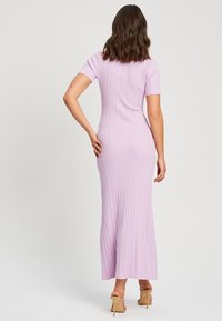 Femme debout, dos tourné vers la caméra, portant une robe longue moulante côtelée lavande et des sandales à talons hauts couleur chair sur un fond clair.