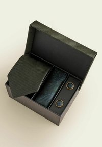 Mörkgrön texturerad slips, matchande näsduk och runda manschettknappar med guldkant prydligt arrangerade i en öppen svart gåvobox.