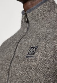 Chaqueta de lana gris con un cuello alto, cremallera completa y un forro interior gris oscuro. Presenta un logo bordado negro en el pecho.