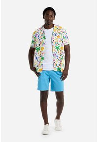 Camicetta a maniche corte, multicolore, con motivi tropicali, abbinata a pantaloni corti blu uni e sneakers bianche. La camicia ha una vestibilità comoda.