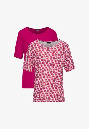 Frau lächelt, trägt ein rosa gemustertes kurzärmeliges Oberteil, weiße gekürzte Hosen und rosa gewebte Schlupfsandalen, steht mit einer Hand in der Tasche.