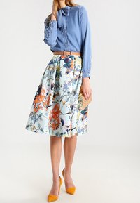 Ljus denimskjorta med fluga, krämfärgad blommig kjol med orange och blå mönster, matchad med gula klackar och en kuvertväska.