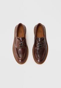 Un paio di scarpe casual in pelle marrone con lacci e suola spessa, viste dall'alto su uno sfondo bianco semplice.