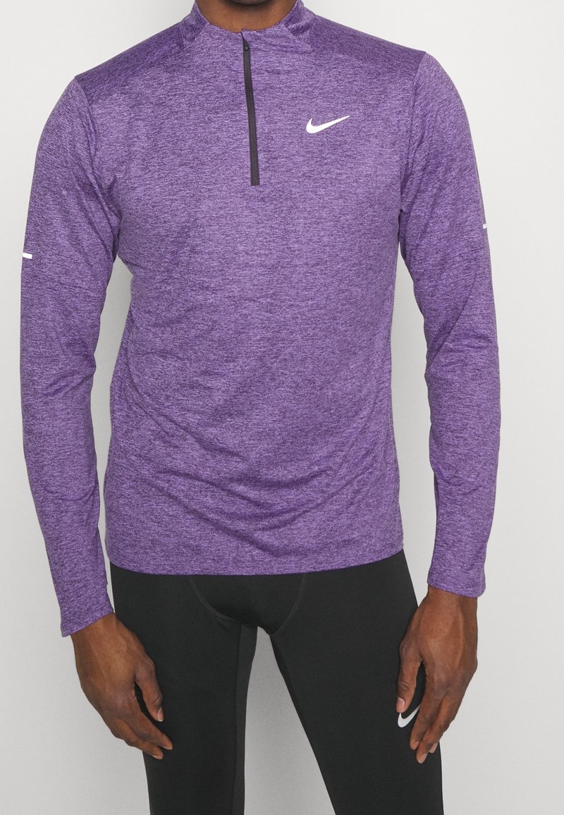 Homme portant un haut Nike violet à manches longues avec fermeture quart de zip et des leggings Nike noirs sur un fond uni.