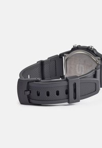 Cinturino in gomma nera con un design testurizzato, numerosi fori per la regolazione e una fibbia in metallo, con il retro dell'orologio in acciaio inossidabile visibile.