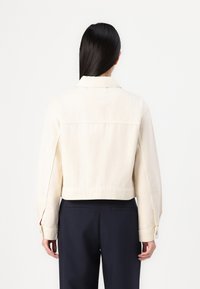 Chaqueta de mezclilla crema recortada con cuello puntiagudo, puños con botones y bolsillos laterales. La vista trasera muestra detalles de costura simples.