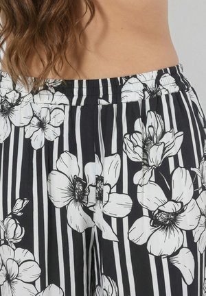 Sort-hvide bukser med lodrette striber og store hvide blomsterprint, båret af en person med langt brunt hår og synlig mave.