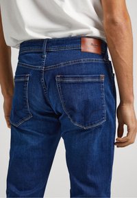 Dark blue denim jeans with a textured surface, featuring back pockets and a leather brand patch on the waistband.