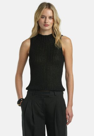 Ženska z blond lasmi, oblečena v črno brezrokavnik iz pletenine in črne hlače z visokim pasom in naborki, stoji pred enobarvnim ozadjem.