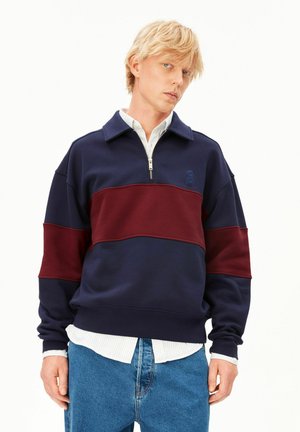 Junger blonder Mann, der einen marineblauen und burgunderfarbenen Quarter-Zip-Sweatshirt über einem weißen Hemd und blauen Jeans vor einem einfarbigen Hintergrund trägt.