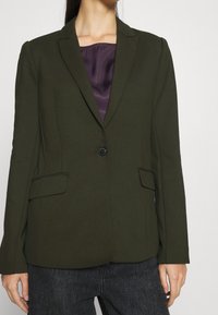 Mujer vistiendo un blazer verde oscuro con un botón abrochado sobre una camiseta morada y jeans oscuros, de pie frente a un fondo neutro.