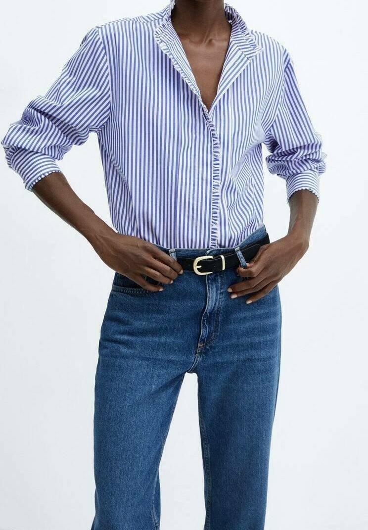 Personne portant une chemise à rayures verticales bleues et blanches avec les manches retroussées, rentrée dans un jean bleu taille haute avec une ceinture noire.