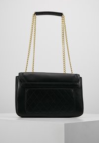 Svart läderaxelväska med kviltad framficka och guldkedjeaxelrem som vilar på vit pelare mot grå bakgrund.