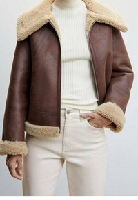 Chaqueta de símil cuero marrón con un cuello y puños de borreguito color crema, cierre de cremallera y un ajuste relajado, combinada con un jersey de cuello alto beige de canalé y vaqueros claros.