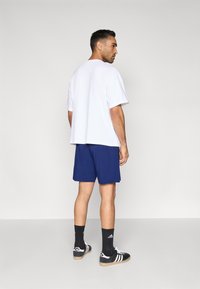 Camiseta branca de algodão, corte solto, combinada com calções azul-marinho. Ténis de desporto a preto e branco, meias pretas com logótipo branco, fundo simples.