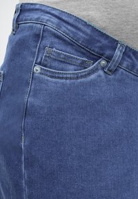 Sinine teksapükste lähedalt vaadatav tasku, kus on nähtavad õmblused, neetide ja vööaasa, mida kantakse halli pealispidisega.