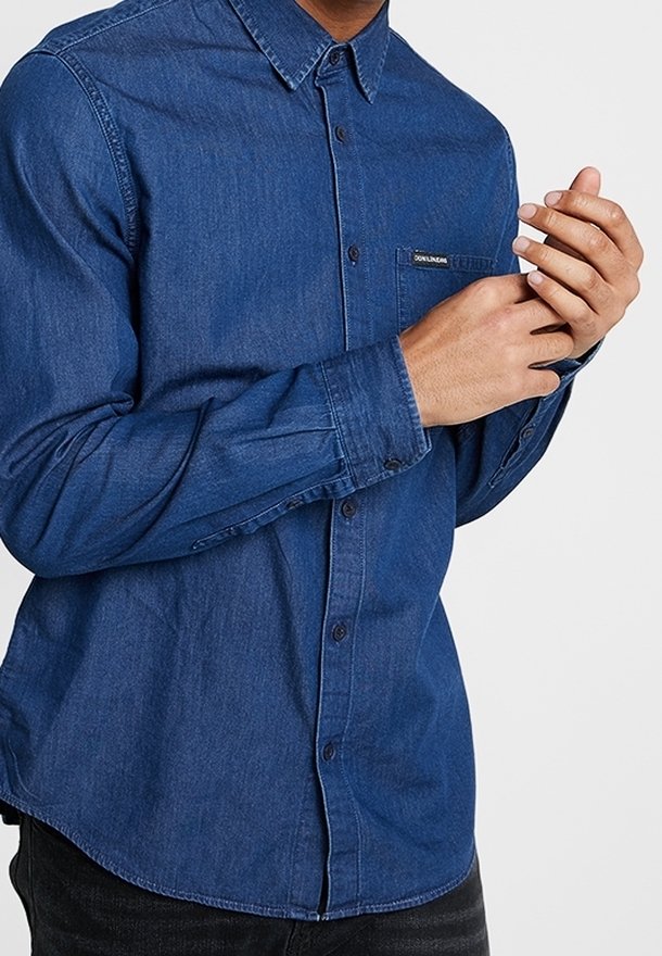 Knapp-up skjorta i mörkblå denim, långa ärmar, framficka och en svängd nederkant. Har subtil söm och en märkesetikett på fickan.