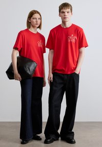 Κόκκινα βαμβακερά μπλουζάκια με το λογότυπο Calvin Klein, συνδυασμένα με σκούρα μπλε τζιν παντελόνια με φαρδιά μπατζάκια. Το μοντέλο στα αριστερά κρατά μια μαύρη δερμάτινη τσάντα.